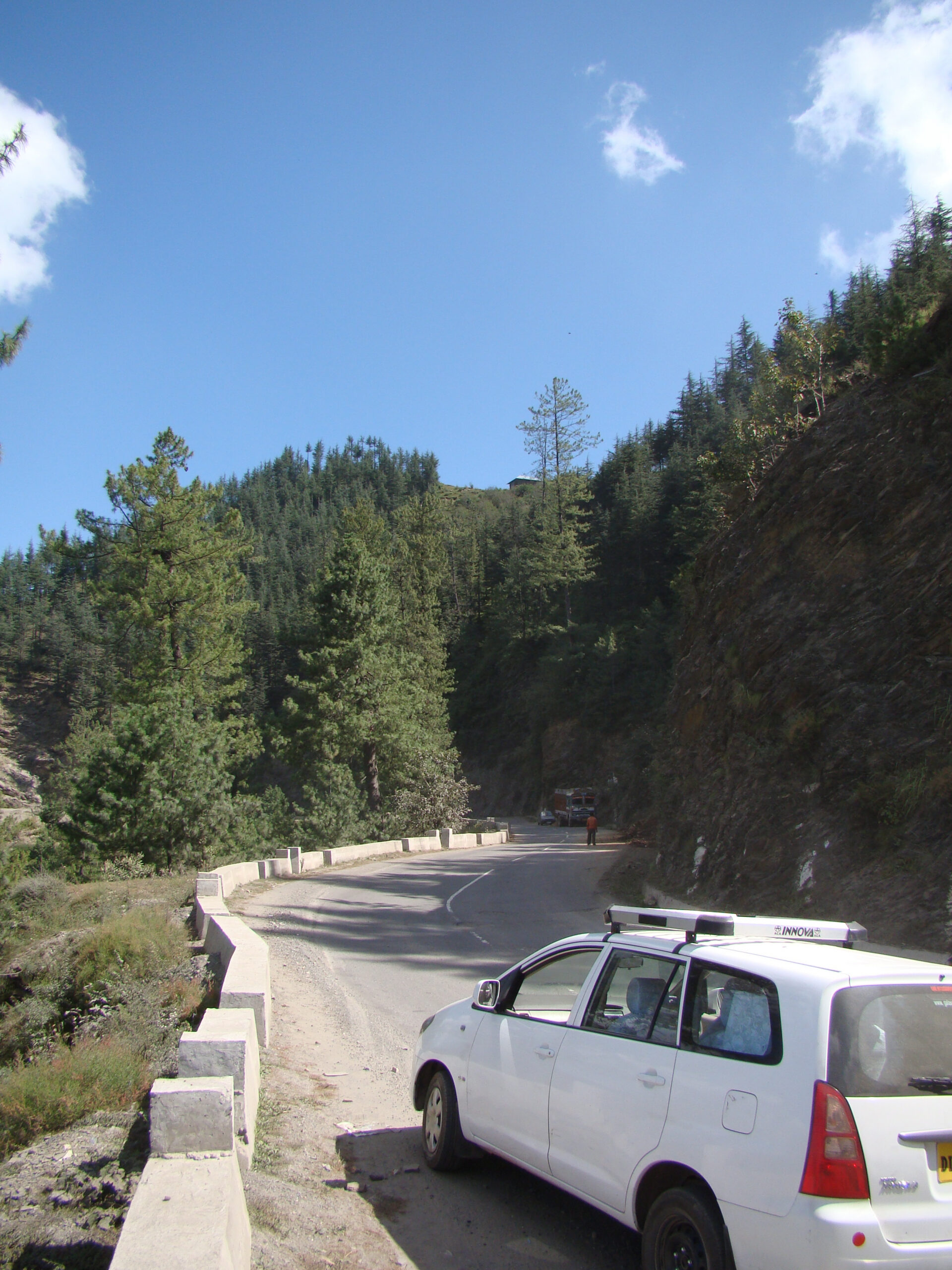 White vehicle parked beside a winding mountain road bordered by pine forests in northern India